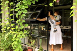 Janela metálica quadriculada preta, sendo aberta por mulher com criança no colo. Vegetação pendente e jardim na frente da janela.