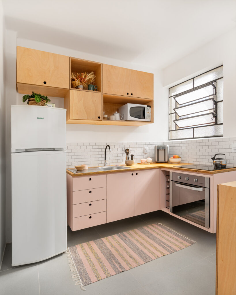 Cozinha com balcão rosa, tampo e aéreo em madeira multilaminada (compensado naval), fogão de indução, revestimento de parede branco e piso em revestimento cinza. Janelas pretas.