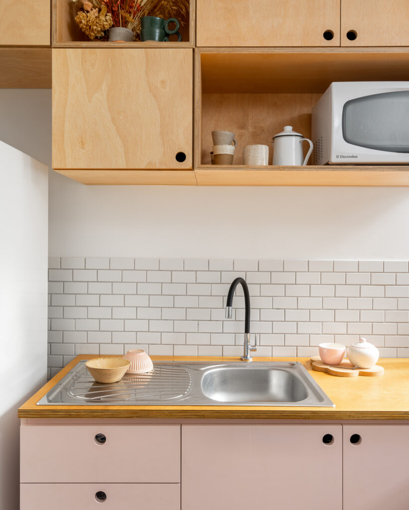 Cozinha com balcão rosa, tampo e aéreo em madeira multilaminada (compensado naval), revestimento de parede branco. Torneira preta e cuba de inox.