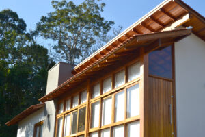 Casa com porta de madeira, janelas de vidro, madeiramento do telhado aparente, paredes brancas. Fundo com vegetação abundante.