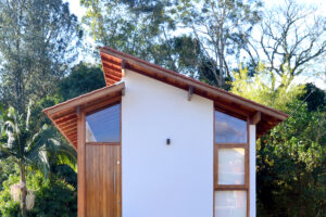 Casa com porta e janela de madeira, madeiramento do telhado aparente, paredes brancas. Fundo com vegetação abundante.